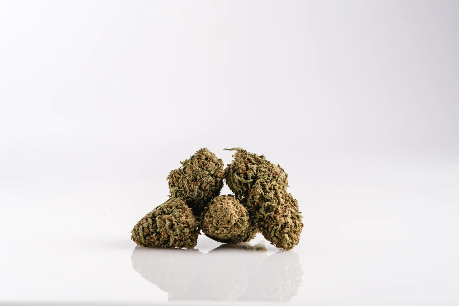 A minimalistic composition of several dried cannabis buds stacked together on a reflective white surface. The neutral background emphasizes the structure and color of the buds. This visual represents the diversity of types of cannabinoids, including minor compounds like CBG and CBN, which play a role in cannabis’s therapeutic benefits.