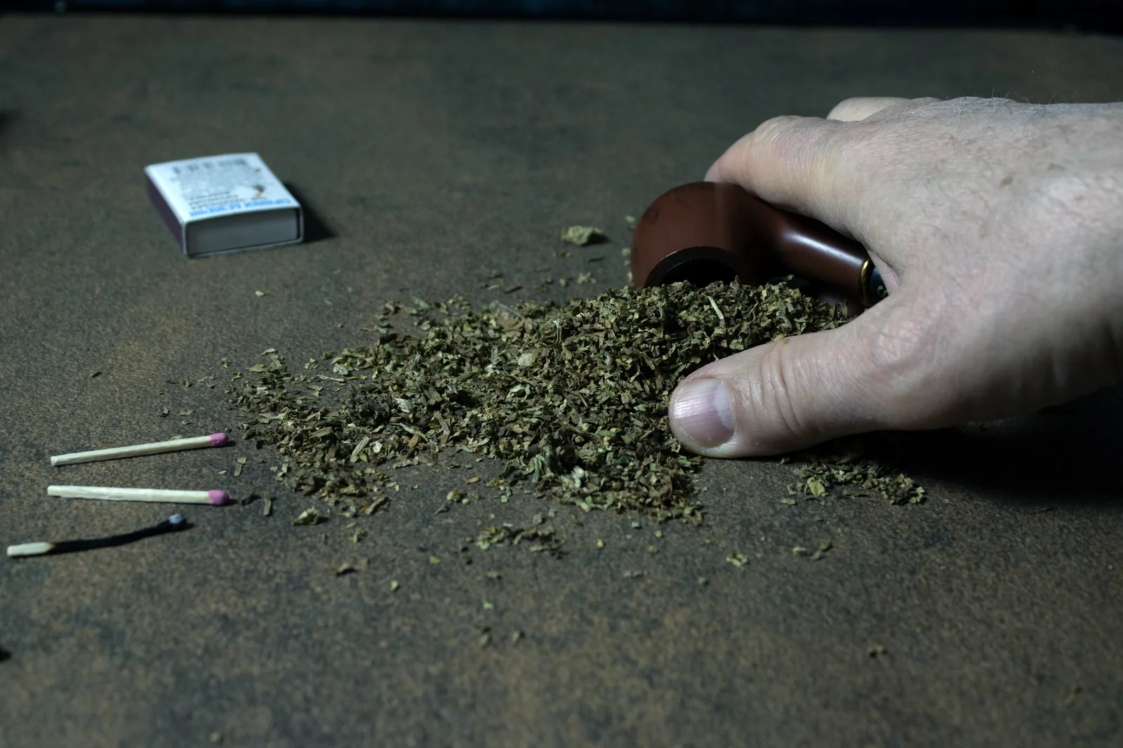 A close-up of a hand holding a wooden smoking pipe next to a pile of dried herbal material, with matches and a matchbox nearby. The scene suggests traditional smoking methods, possibly involving different types of marijuana or tobacco blends.