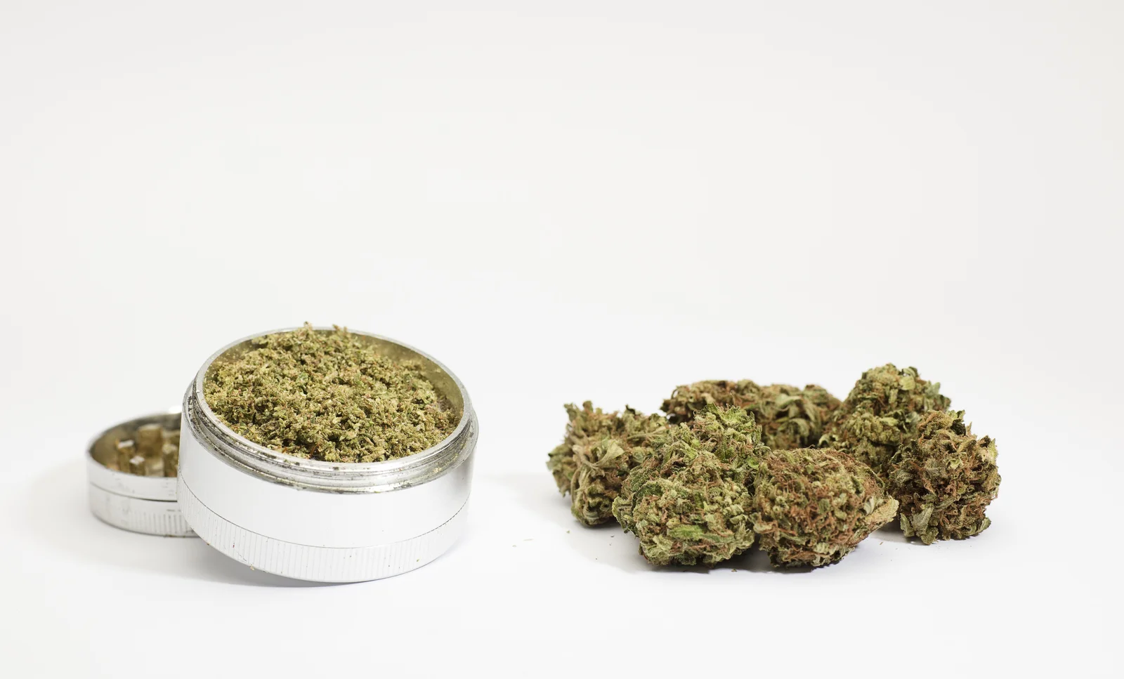A close-up image of a metal grinder filled with finely ground cannabis next to dense green buds on a clean white background. This minimalistic setup reflects product consistency and premium quality, as seen in the Top 10 cannabis Dispensaries in New Jersey.