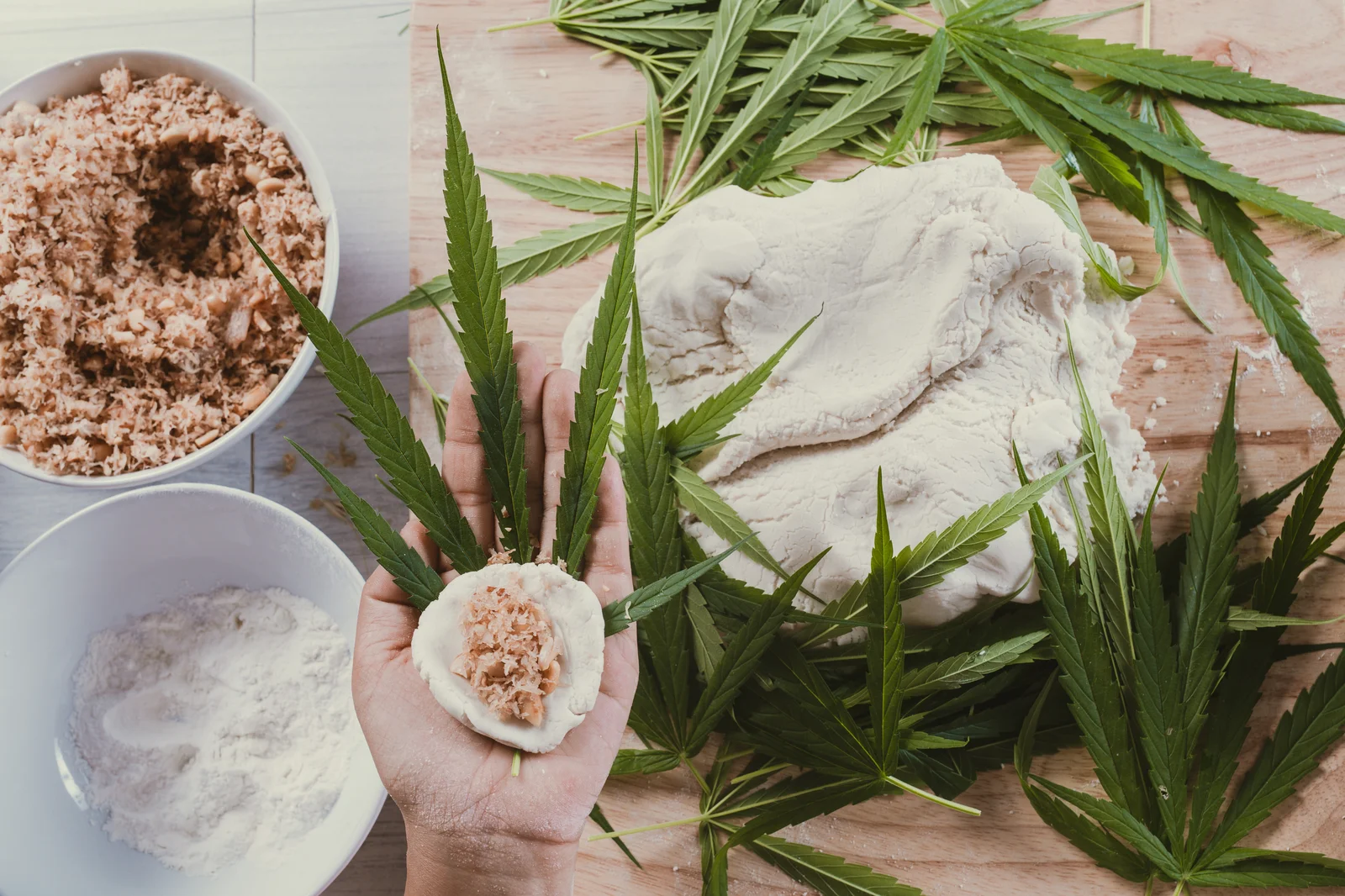Hand holding raw dough and shredded filling on top of a cannabis leaf, surrounded by various ingredients and different types of marijuana leaves on a wooden surface.