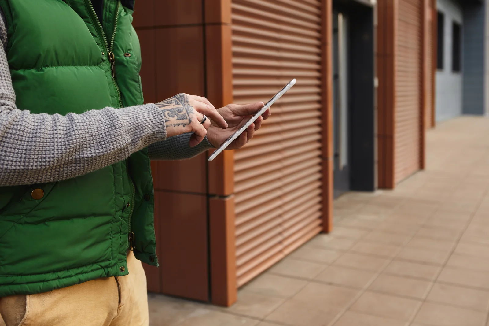 A close-up of a man with a tattooed hand using a tablet outdoors near a commercial building, possibly managing data related to how to grow a cannabis plant from seed through digital tools.