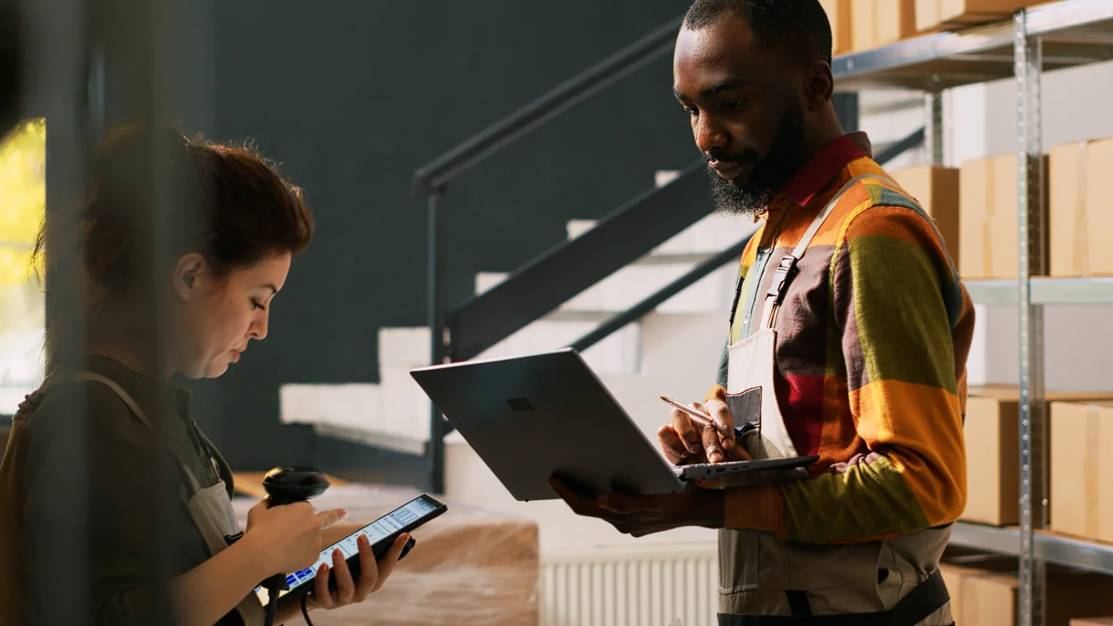Two warehouse workers examine inventory using tablets and laptops in front of shelves, highlighting logistics and tracking systems important in how to grow a cannabis plant from seed and bring it to market.