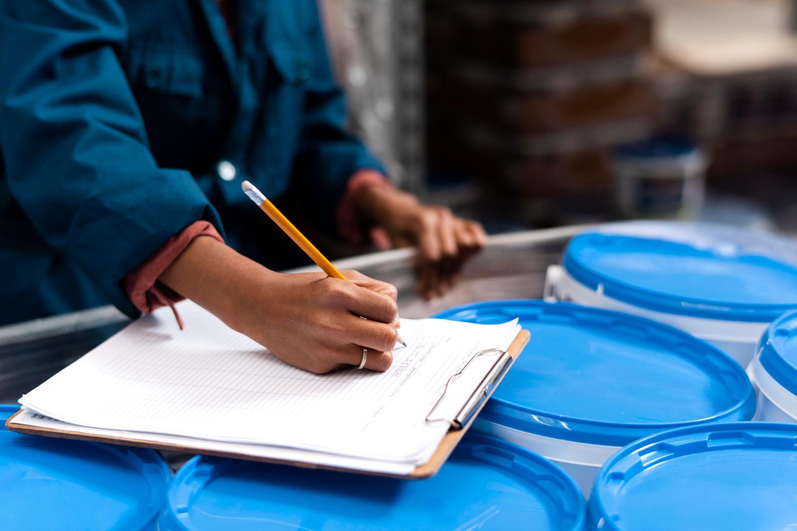 A person in a blue jacket writes on a clipboard near blue-lidded containers in a facility setting. This could represent part of the post-harvest tracking or processing phase, helping to answer questions like how do I know when to harvest cannabis by monitoring quality benchmarks and timelines.