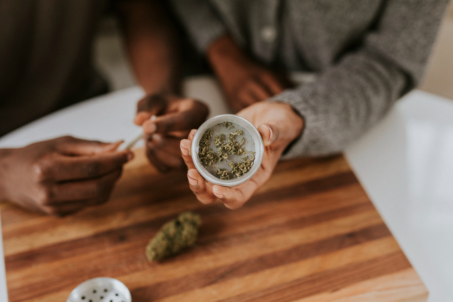 How to grow CBD flower at home illustrated by Hands holding an open metal grinder filled with ground cannabis, with a joint being rolled on a wooden board in the background