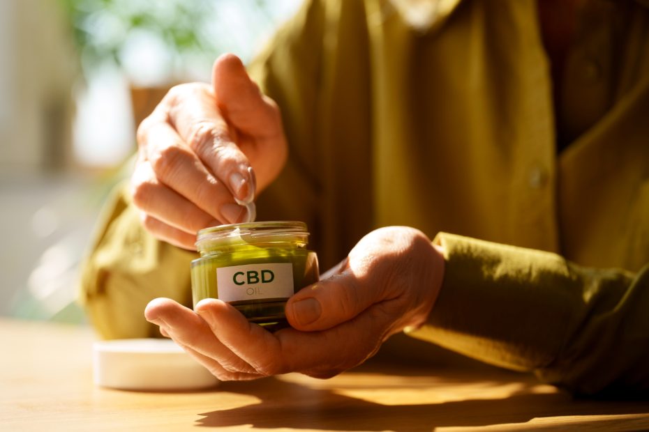Close-up of hands applying a green topical balm from a small glass jar labeled 'CBD Oil' to the skin, illustrating the use of CBD topicals after learning how to grow CBD flower at home